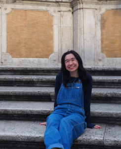A smiling young woman wearing a black long sleeved shirt and blue overalls. She is sitting on stone steps with a white and tan marble wall at the top.