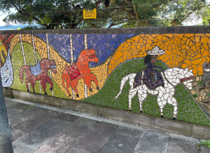 A colorful mosaic mural with three horses (brown, red, white). The one in the lead, on the right, has a person with a sombrero riding. The other two horses look like they are part of a carousel, possibly.